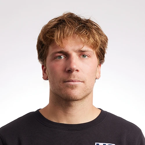 Person wearing a black crewneck shirt with a small logo, standing against a plain light gray background.