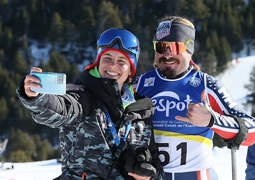 Two people in ski gear take a selfie on a snowy slope, one wearing a race bib with Espot and number 51, trees in the background.