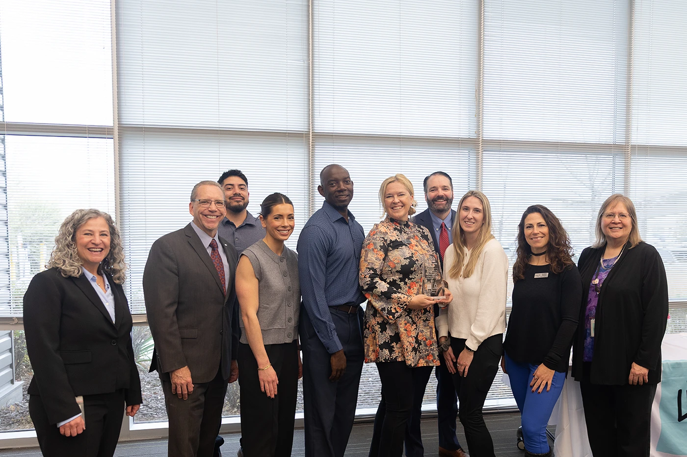 A group of eleven people stand together in a bright indoor space with large windows behind them. They are posed for a group photo, with several individuals in the center holding an award or trophy. A table covered with a white cloth and a teal banner reading “Live On.” is visible to the right. The group is dressed in a mix of business and business‑casual attire.