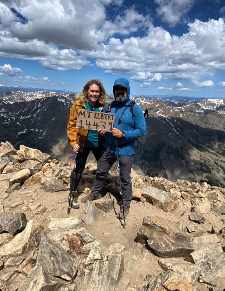 A woman and a man standing on the rocky summit of a mountain, holding a wooden sign that reads MT. ELBERT 14439. They are wearing hiking gear with scenic mountain ranges and a cloudy blue sky in the background.