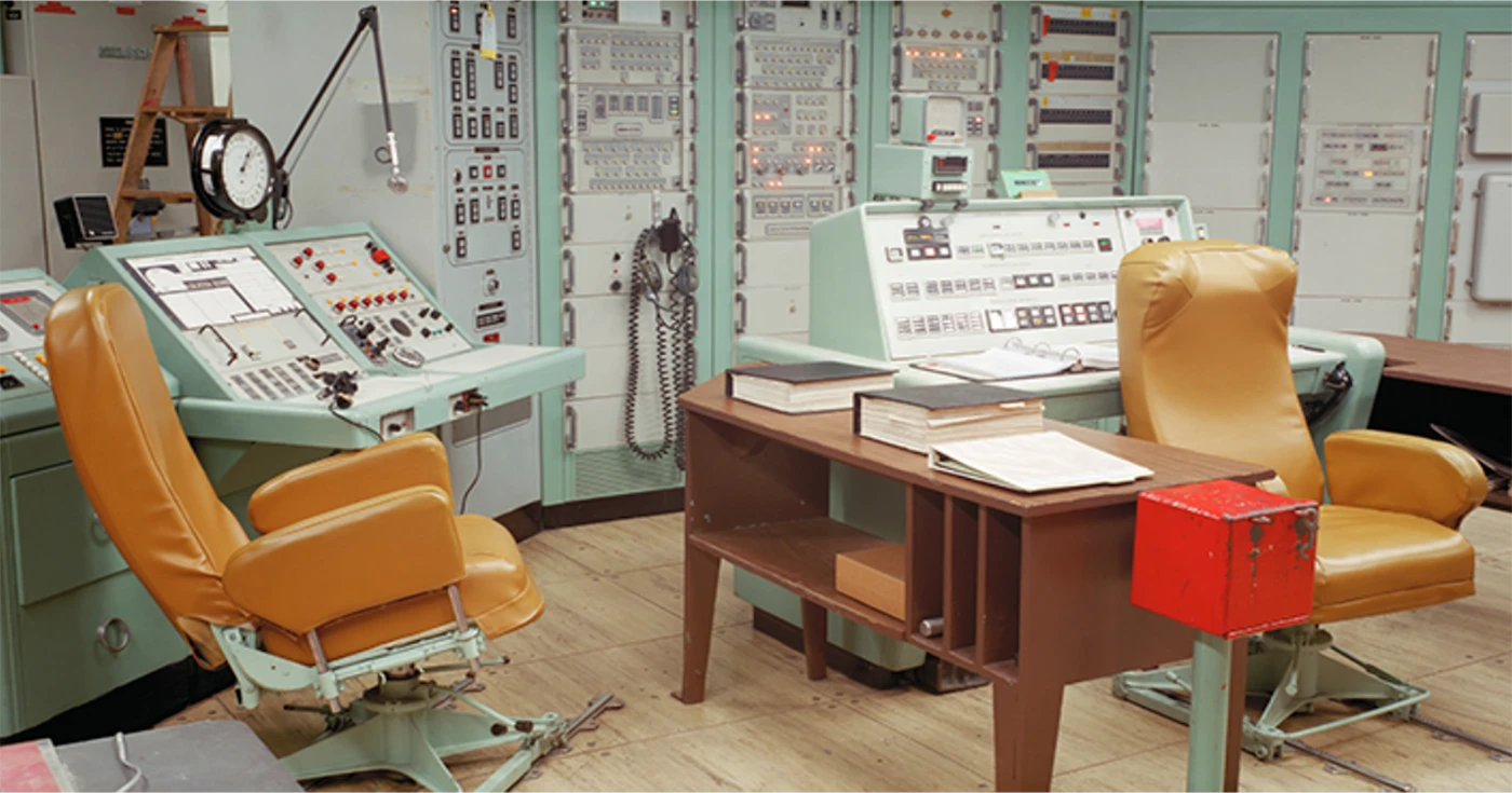 An abandoned office space in a very technical facility. The background is a wall of control panels. The foreground is two wooden desks flanked by green control banks and two orange chairs.