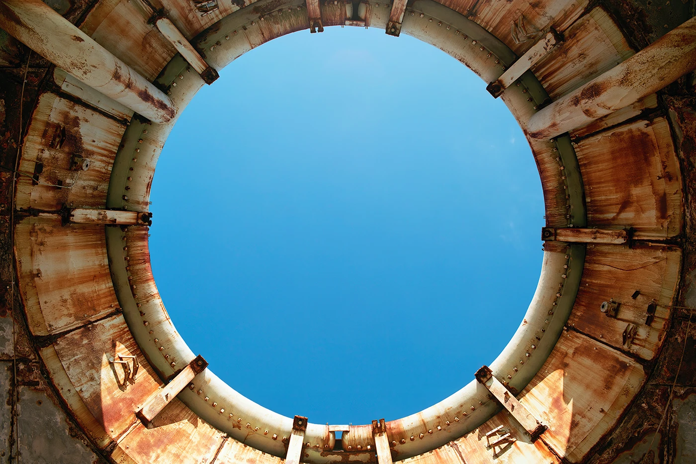 A missile or launch silo seen from below and looking up through the launch aperture. The facility is made from a heavily corroded metal, an off-white color streaked with rust red. The sky beyond is a starkly contrasting blue that fades from more saturated at the bottom to less saturated at the top.