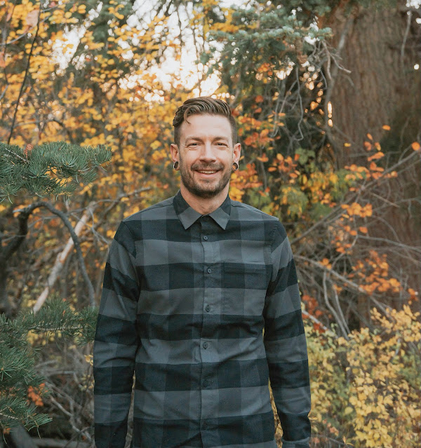 A smiling man in a dark plaid button-up shirt stands outdoors in front of colorful autumn foliage, with pine branches and bare trees in the background.