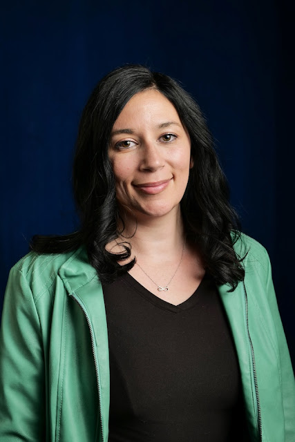 Person with long, dark hair wearing a green jacket over a black top, standing against a dark blue background.