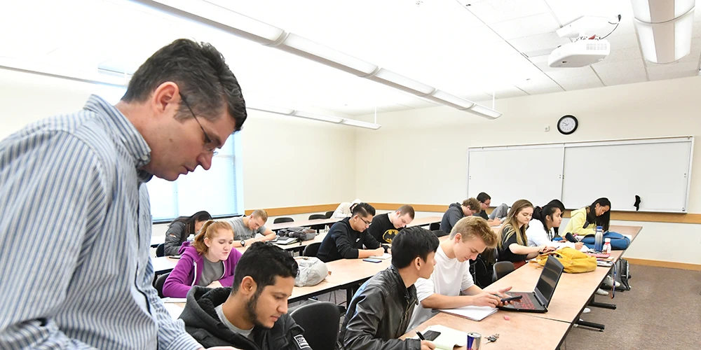 A classroom filled with students working at long tables, some using laptops and others writing in notebooks. A person in the foreground is standing and looking down at a desk. The room is brightly lit with whiteboards, a clock, and a ceiling-mounted projector visible.