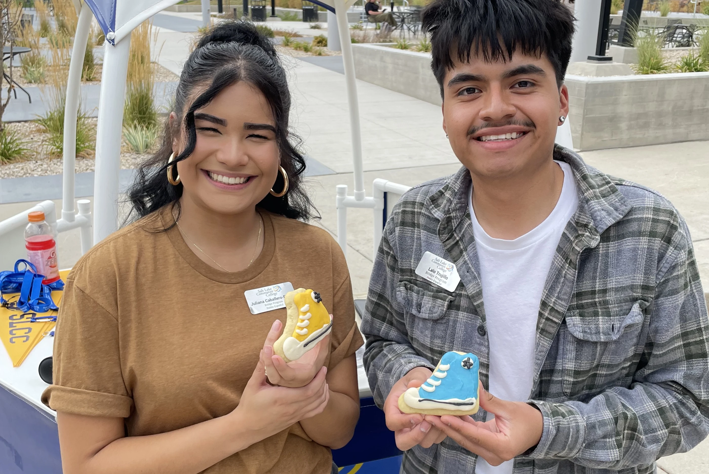 Two students smiling and holding cookies shaped like Converse Chuck Taylor shoes. The cookies are iced with bright icing, one yellow and one blue.