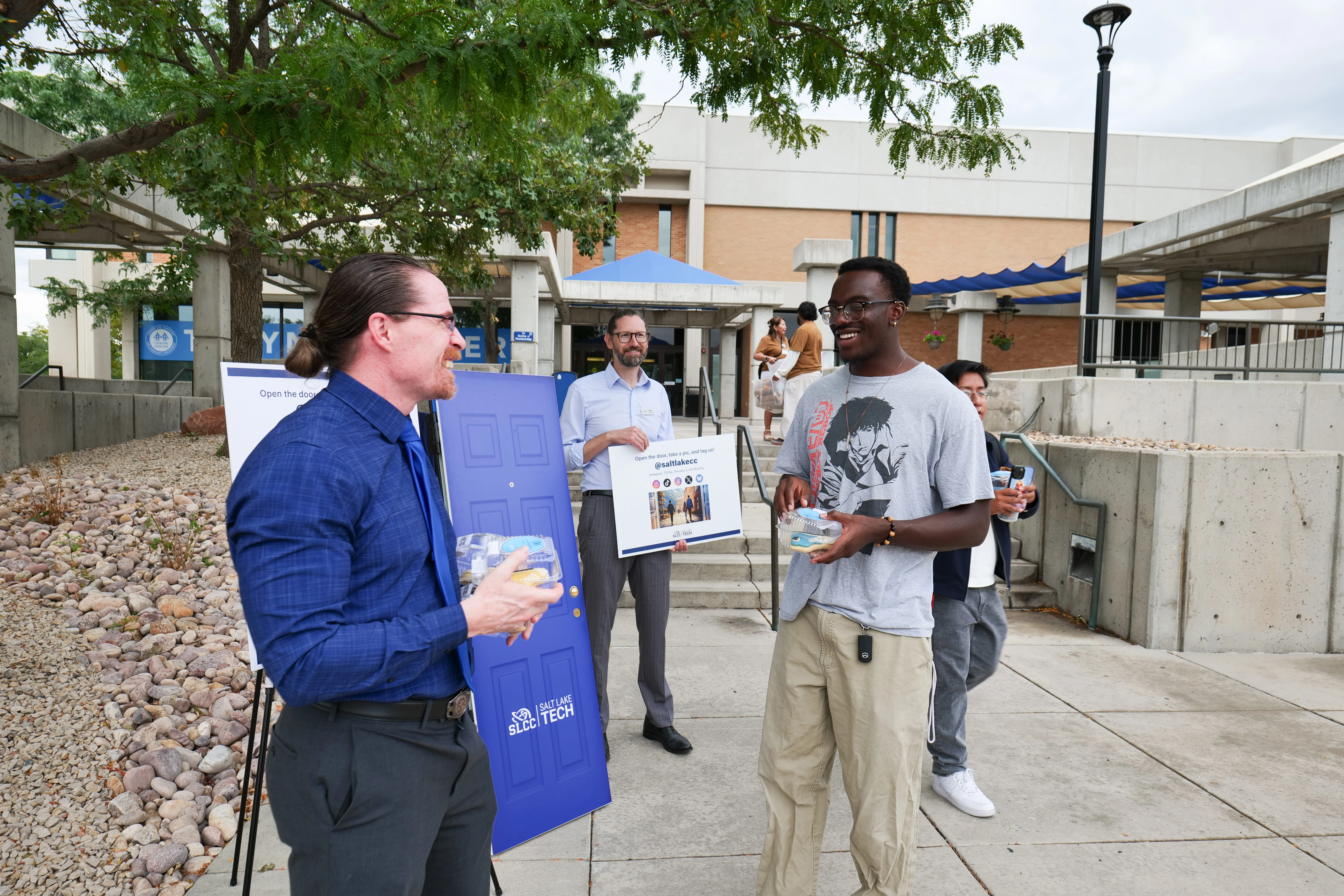 President Greg Peterson, wearing a blue shirt and tie, smiles while speaking with a student in a grey anime shirt. Both are holding cookies in plastic clamshell containers.
