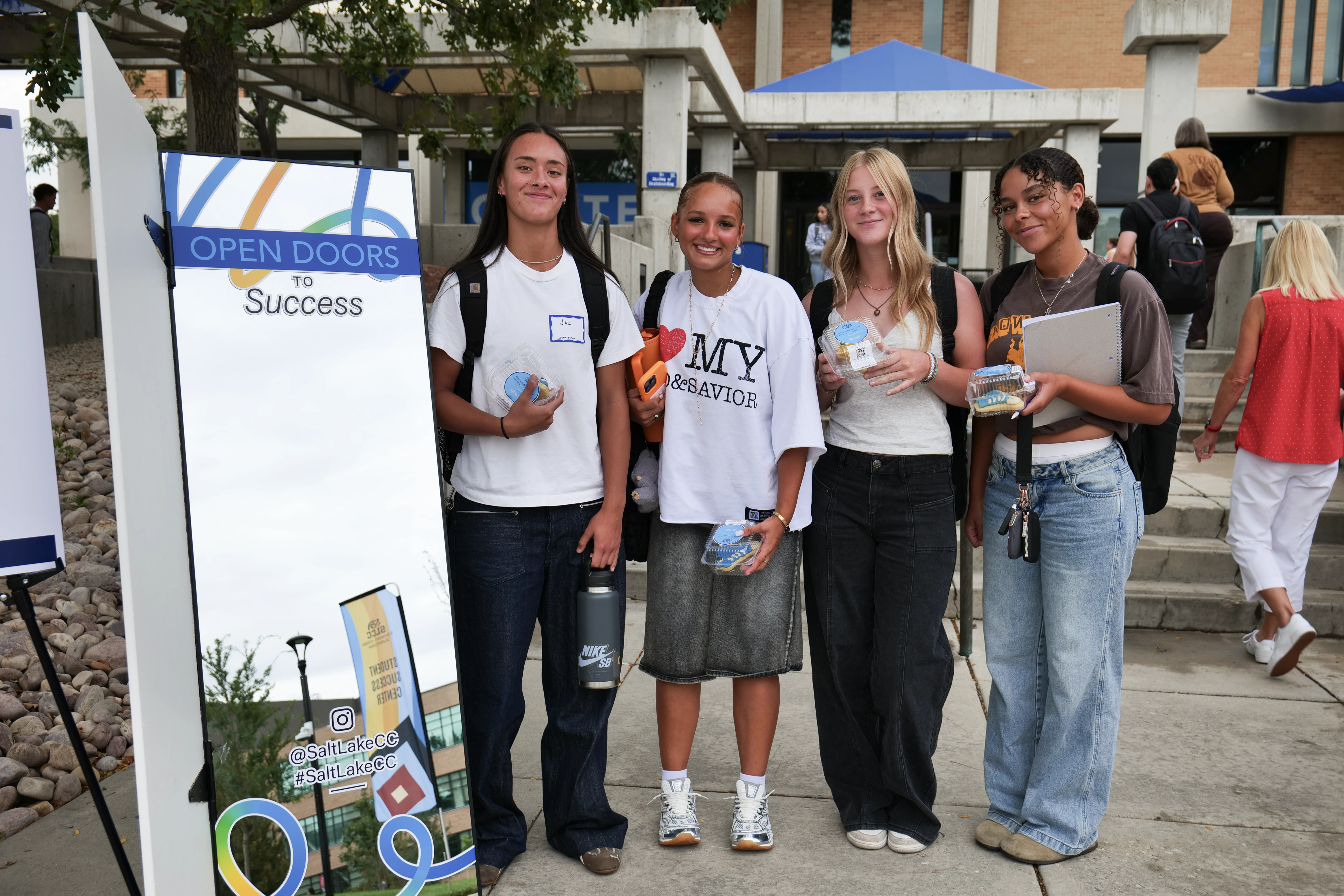 Four students stand holding bags, notebooks, and cookies in plastic clamshells. Next to them is a mirror with vinyl sticker lettering on it that reads: Open Doors to Success.