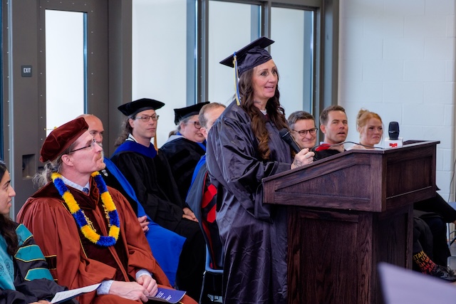 Graduation ceremony with a person in cap and gown standing at a podium giving a speech. Several individuals in academic regalia are seated behind the speaker.