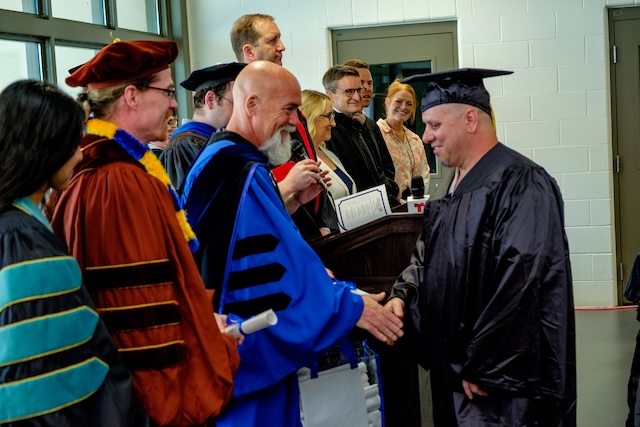 Graduate in black cap and gown shaking hands with an individual in academic regalia during a graduation ceremony. Several others in academic regalia are present, with some standing behind a podium.