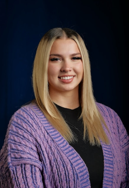 Person with long, straight blonde hair wearing a purple knitted cardigan over a black top, standing against a dark blue background.