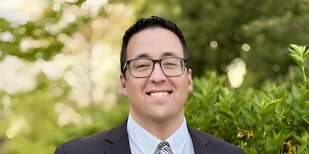 Gary Verón, the new Athletic Director of Salt Lake Community College, smiles at the camera in a professional outdoor headshot. He is wearing glasses, a dark business suit with white dress shirt and diagonal striped tie, standing against a backdrop of green foliage.
