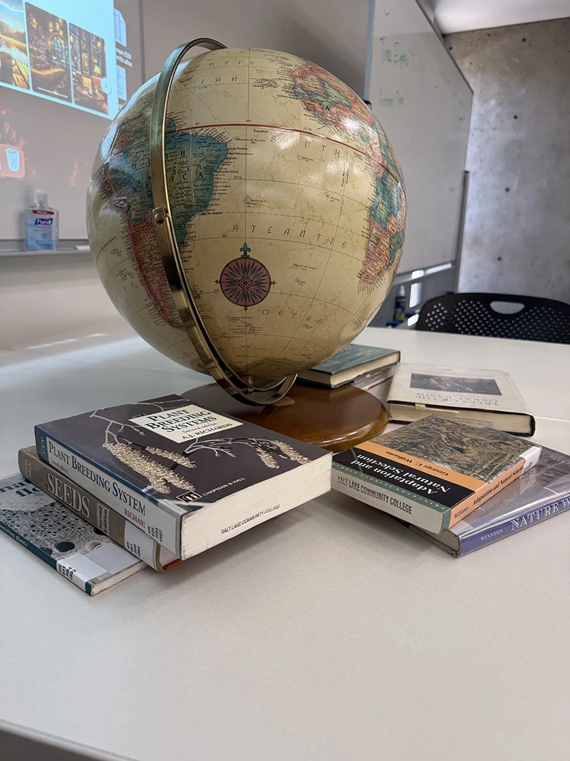 A classroom table with a globe surrounded by books on topics such as plant breeding, seeds, and natural history, with a whiteboard and projected screen in the background.