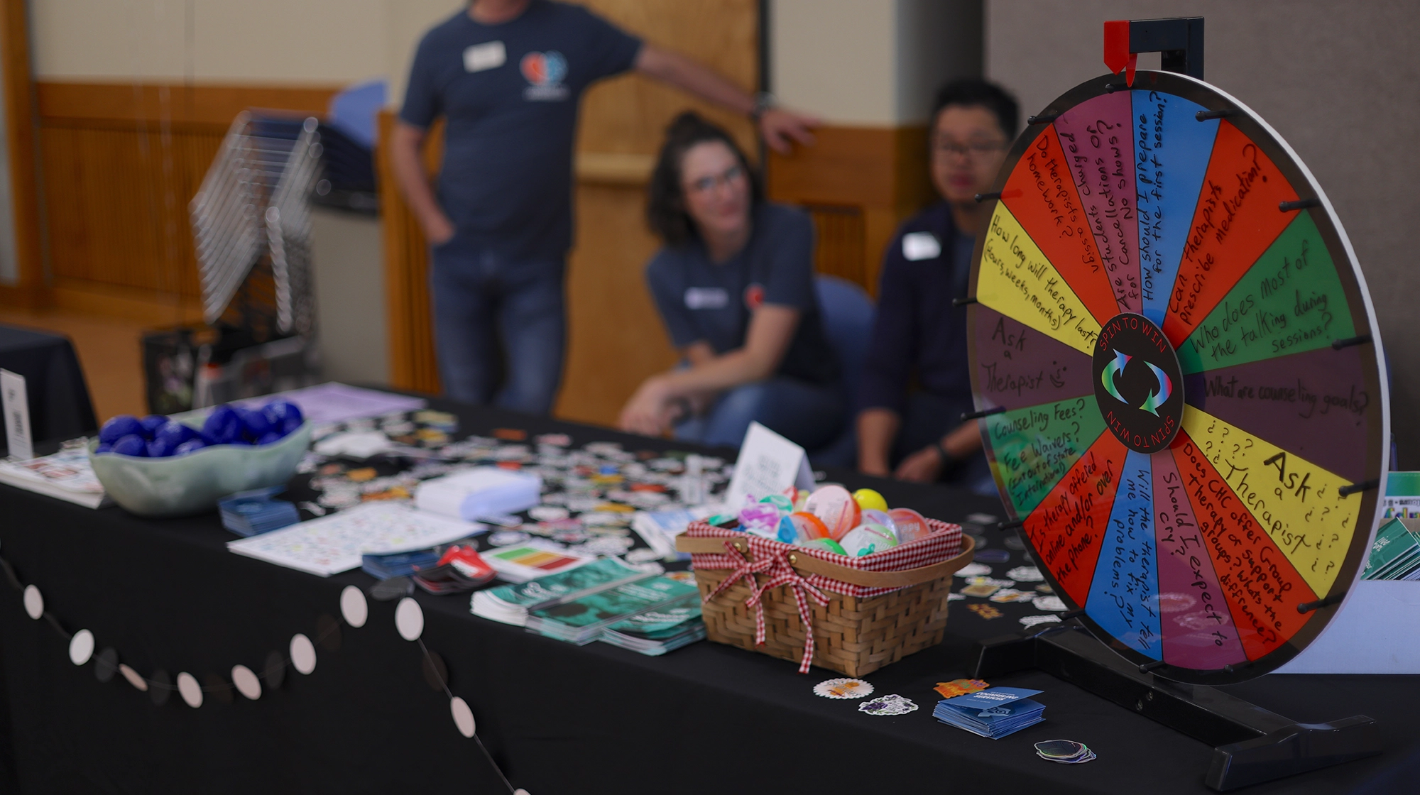 A table with a black tablecloth holding brochures about health resources and a spinning wheel with different resources listed. In the background, representatives from the Center for Health and Counseling are blurry but visible in blue t-shirts.