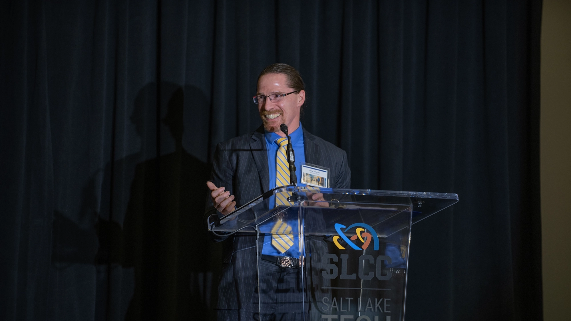 Dr. Greg Peterson stands behind a clear plexiglass lectern in a striped suit wearing a blue shirt and yellow striped tie. He’s gesturing and smiling to someone sitting to the side of the stage.    