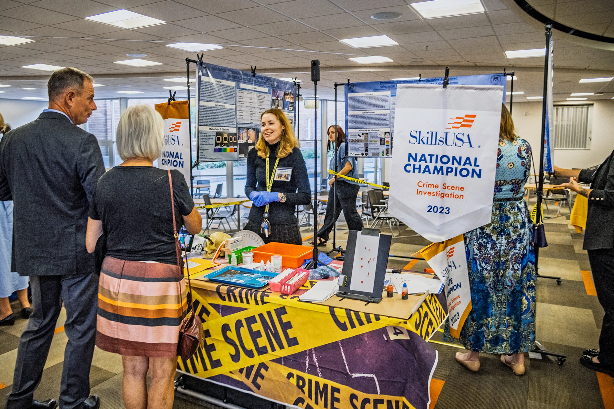 A crime scene demonstration set up by SLCC students and faculty. Event attendees in formal wear are speaking to the interactive exhibit’s attendant. Yellow tape reading "crime scene" is everywhere. 