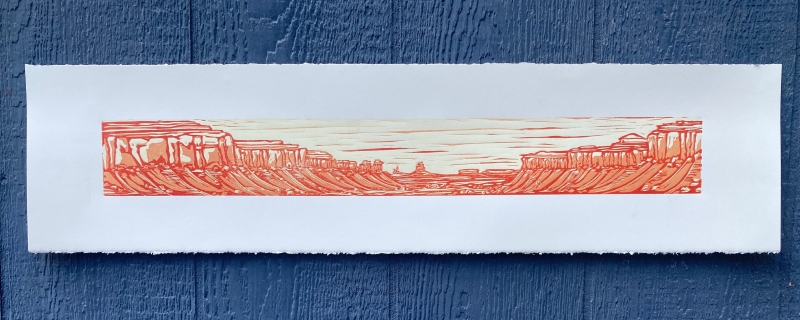 A long, narrow woodcut pressing of a Canyonlands landscape. The background is white paper, roughly torn at the top and bottom edges, and the landscape is printed in cream, orange, and red. Several inches of stark white space surround the print.