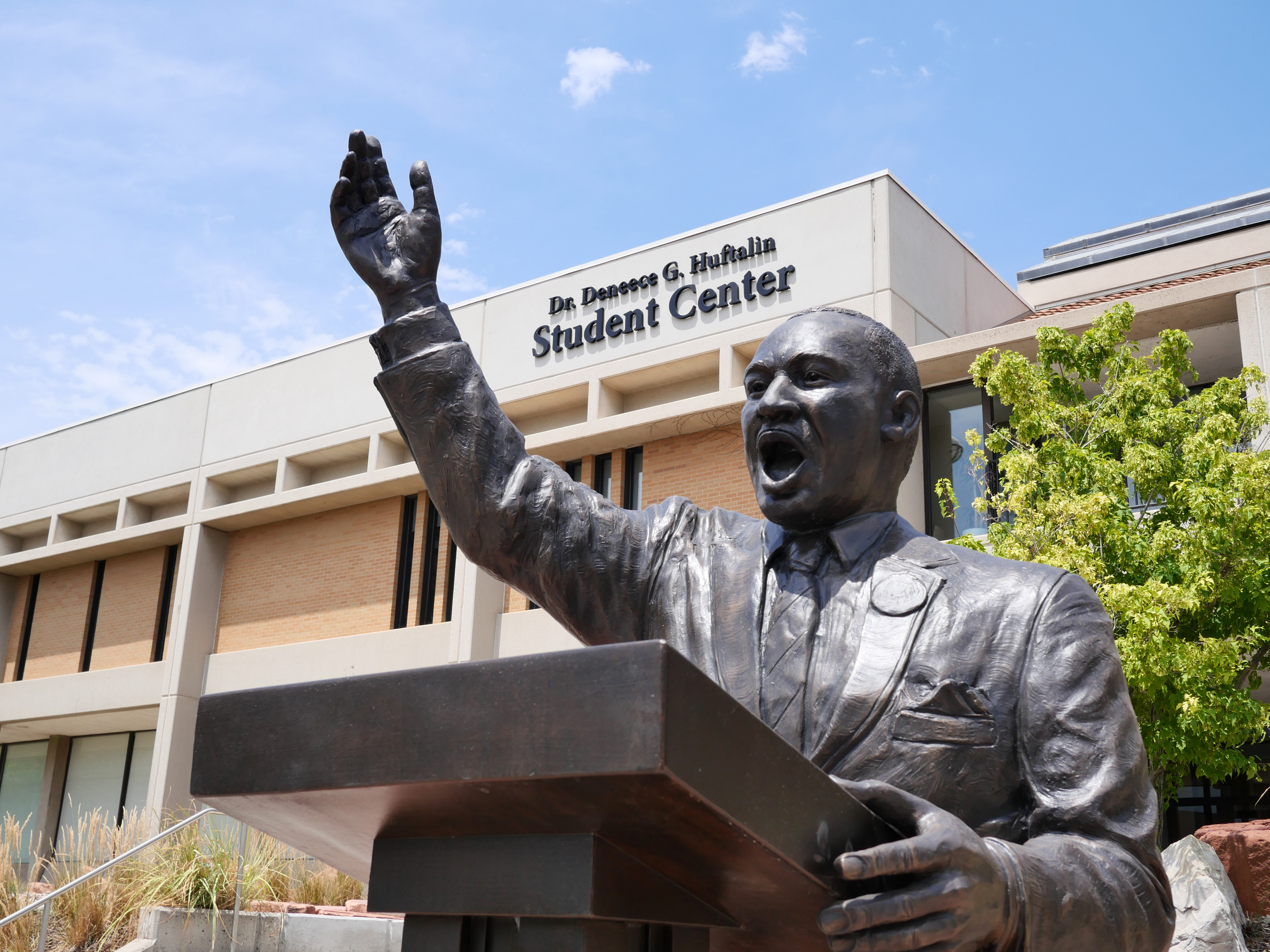 A bronze statue of Dr. Martin Luther King Jr. standing behind a podium with one arm raised, in front of the Dr. Denise G. Huftalin Student Center. The building is modern with large windows, and a tree is visible to the right under a blue sky.