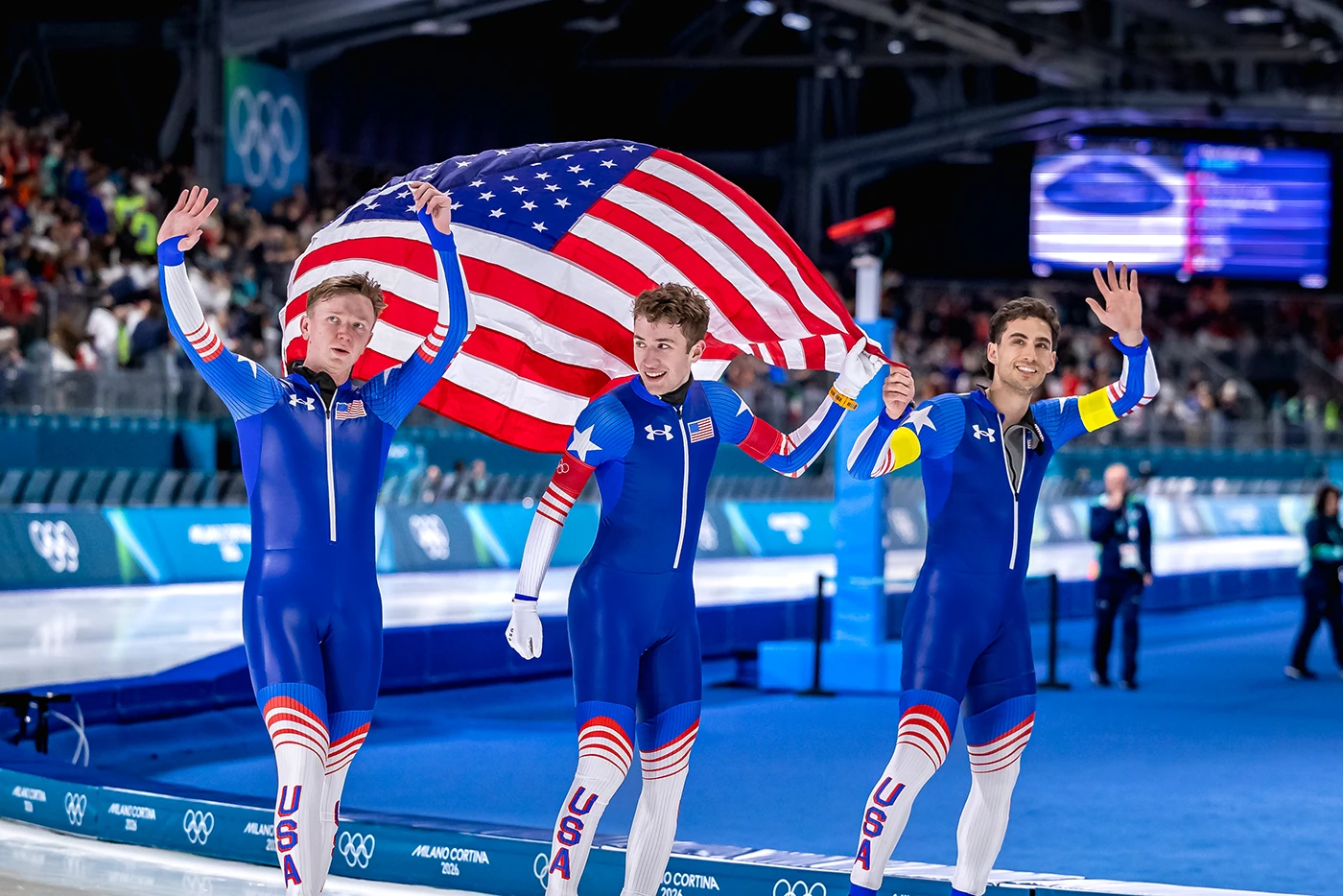 Three speed skaters are rounding a bend wearing blue and white USA skin suits. They're all wearing white helmets with the US flag displayed on the forehead. 