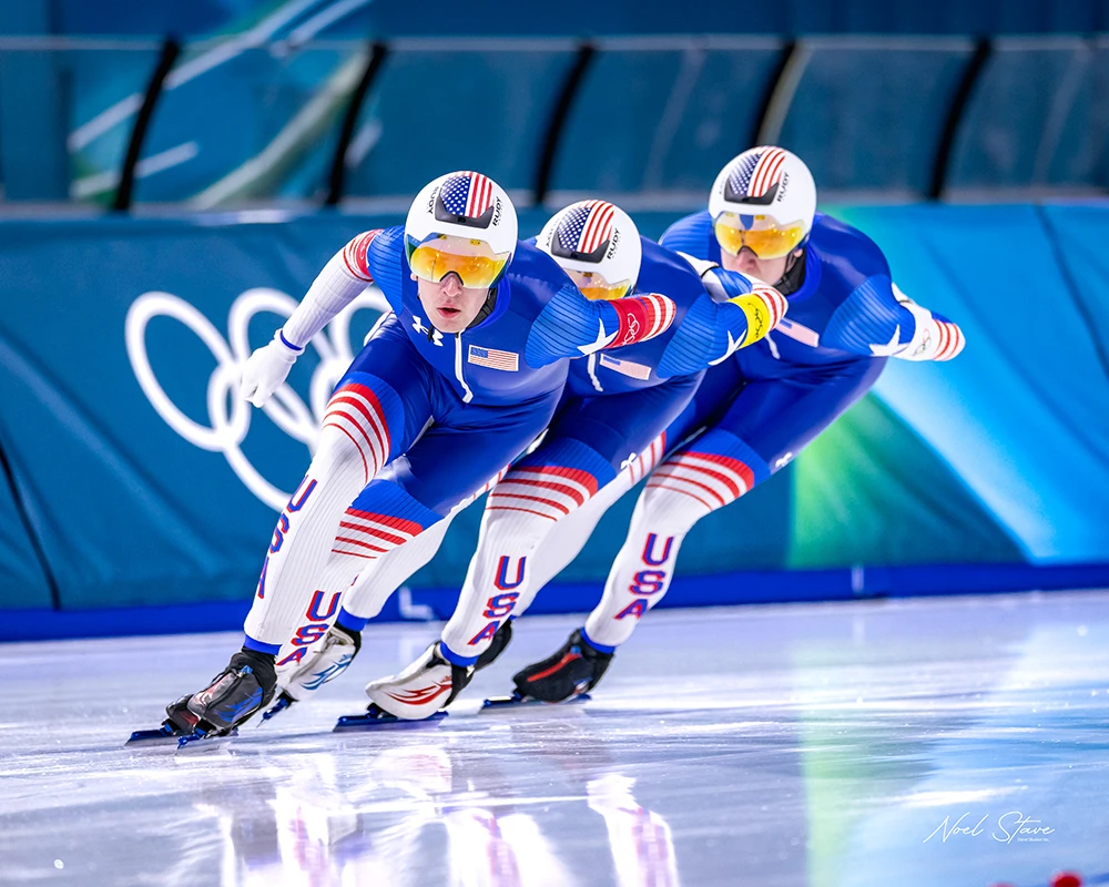 Three speed skaters are rounding a bend wearing blue and white USA skin suits. They're all wearing white helmets with the US flag displayed on the forehead.