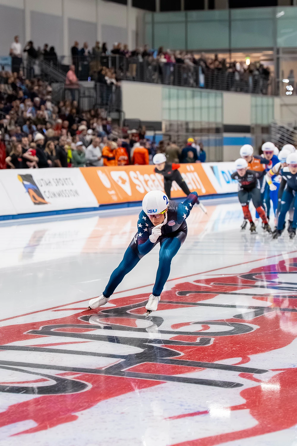 A speed skater with a gap off the front of a chasing group, tucked in an aero position and wearing a dark blue USA skin suit with a white helmet showing the number two.