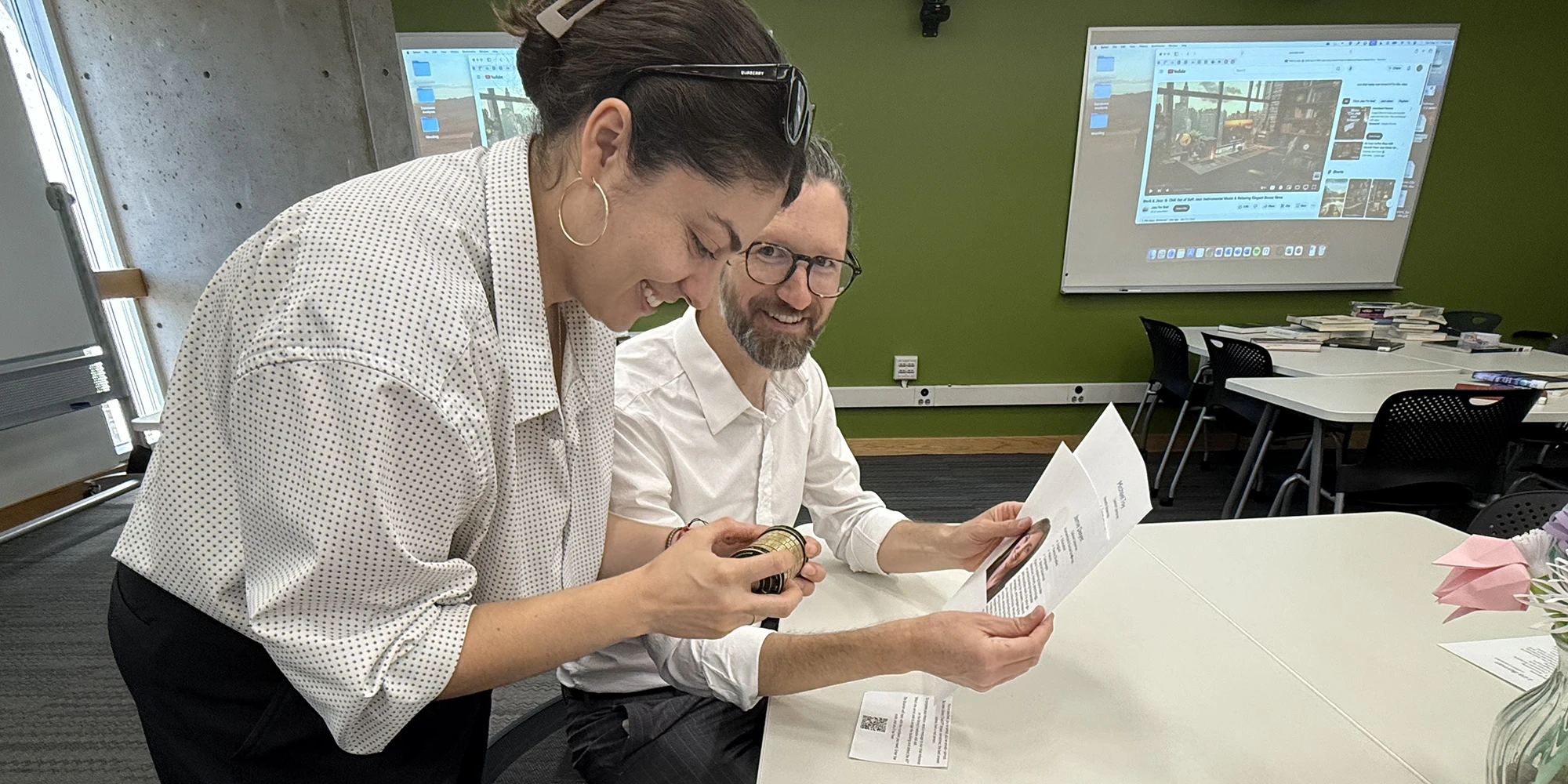 Two people smiling while looking at a small object together at a table, with printed papers spread out in front of them and a projector screen displaying an image in the background.