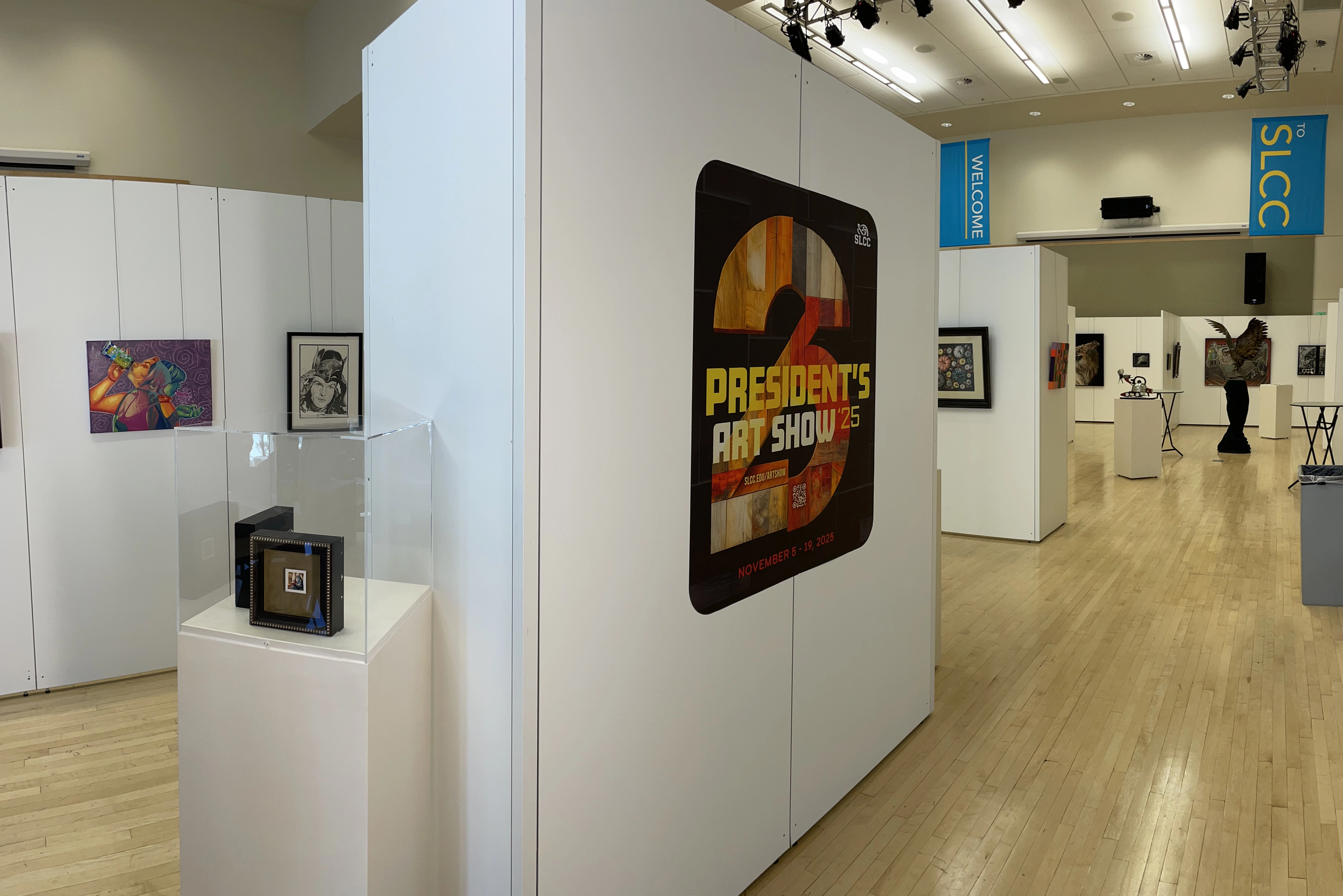 Art gallery with white walls, wood floors, and various paintings and sculptures; poster reads Presidents Art Show 25.
