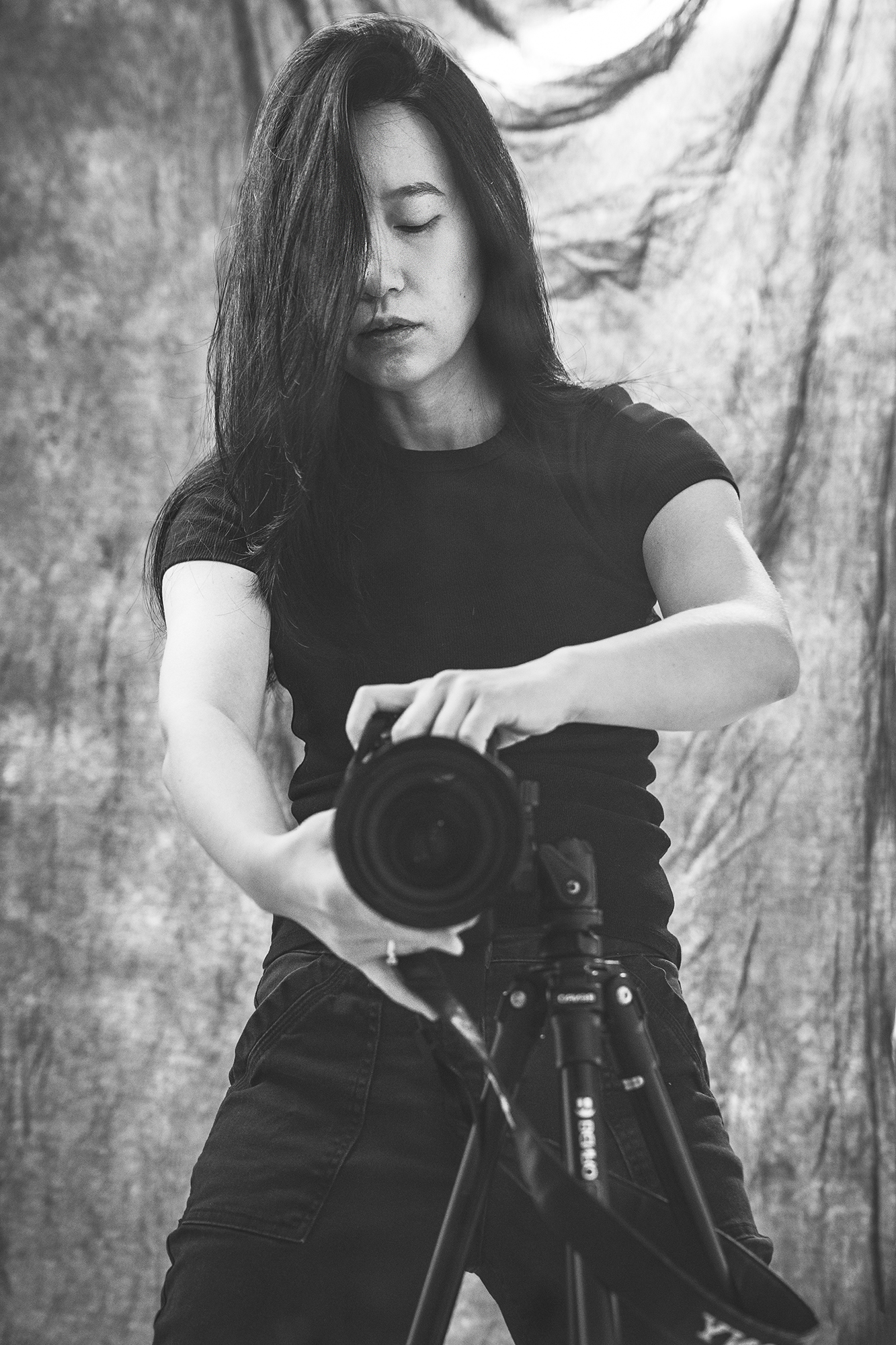 A black-and-white self portrait of the artist wearing dark clothing and pointing a camera into a mirror.