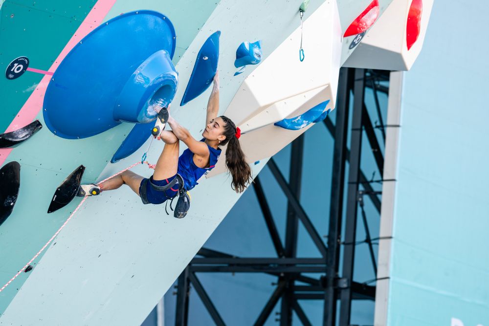 Person climbing an indoor rock wall, wearing harnesses and safety gear, with colorful climbing holds visible on the wall.
