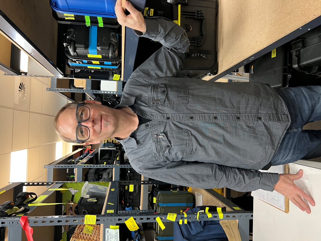 The artist, smiling in matte grey eye glasses and a grey button-up shirt. He's surrounded by shelves of photo and video equipment in large, hardcase storage boxes.