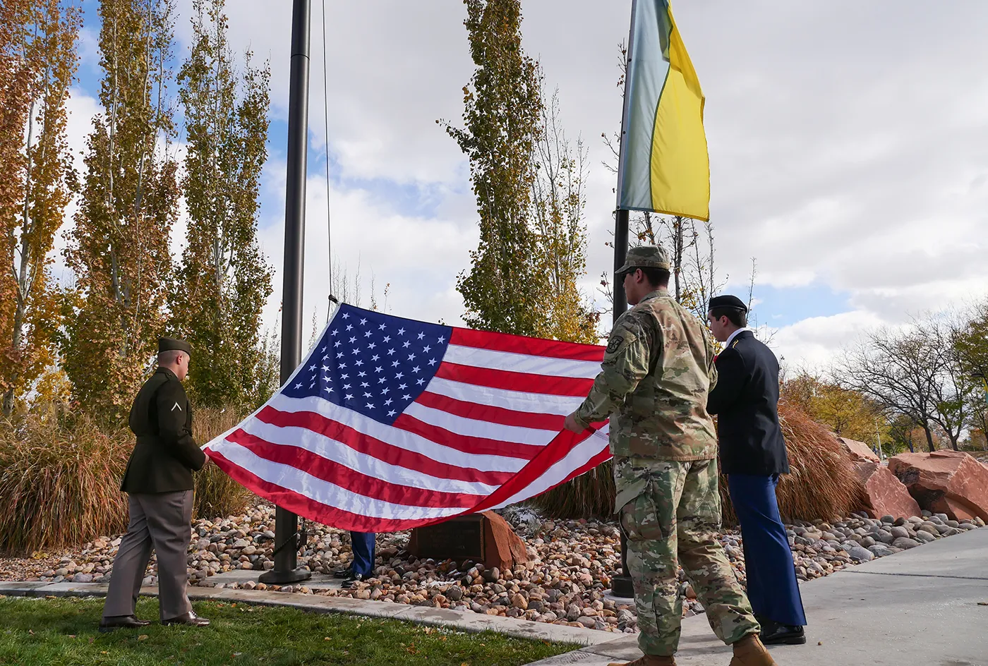 Three service members in military uniforms stand at the base of a flagpole, holding an American flag horizontally during an outdoor Veterans Day ceremony at Salt Lake Community College, November 10, 2022. A SLCC yellow and blue flag flies above them, set against a background of tall, autumnal trees and a partly cloudy sky.