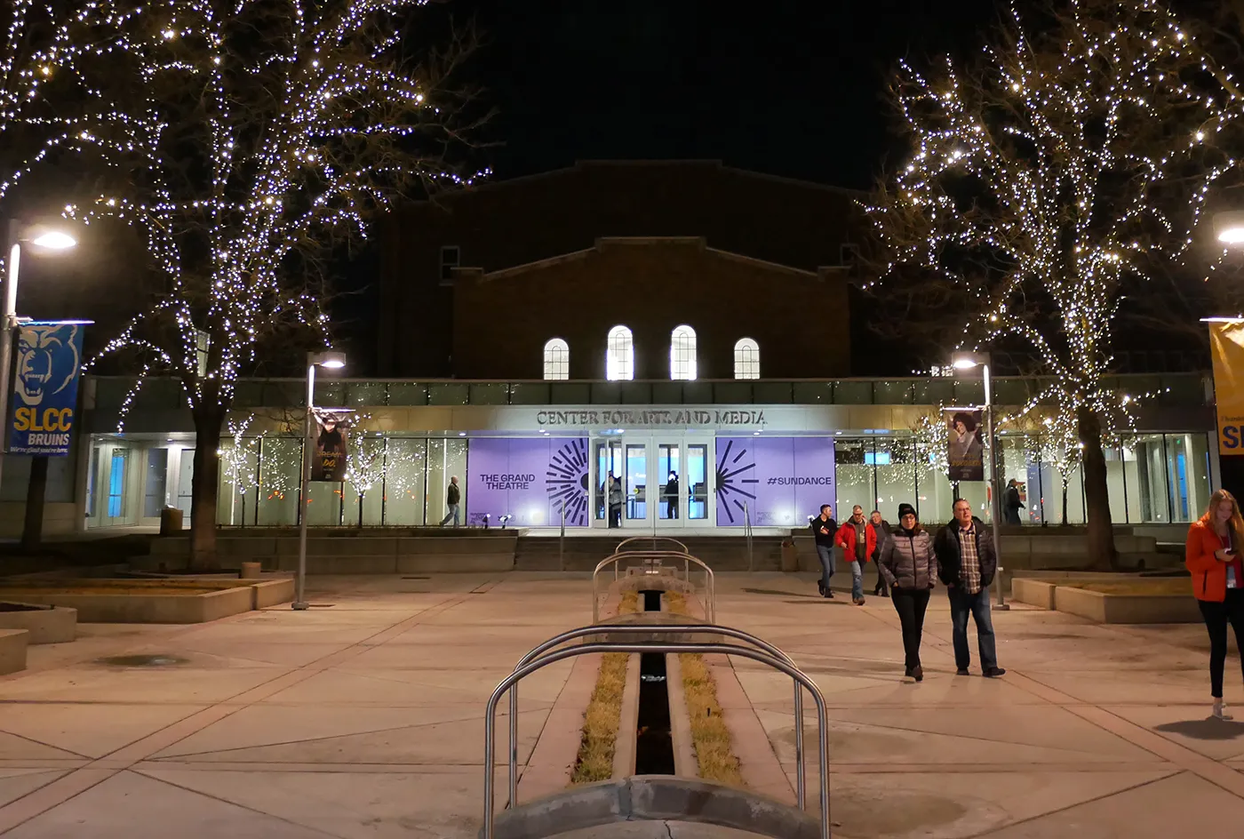Night plaza with lit trees and people walking toward a glass-fronted Center for Arts and Media with Sundance banners.